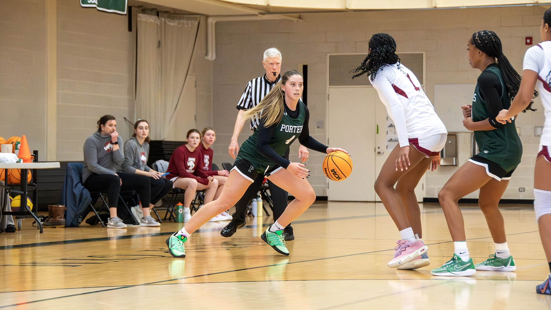 A basketball player in a green uniform dribbles past two defenders on an indoor court as a referee and teammates watch from the sideline.