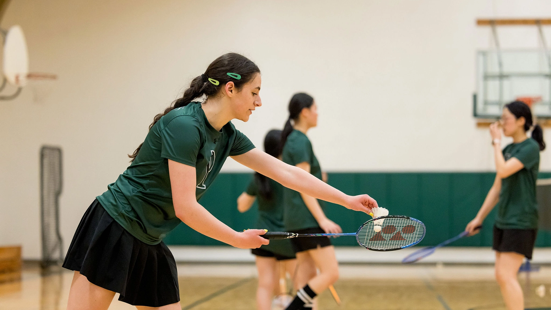 A young woman in a green shirt and black skirt prepares to serve in a badminton game, with three other players in the background on an indoor court.