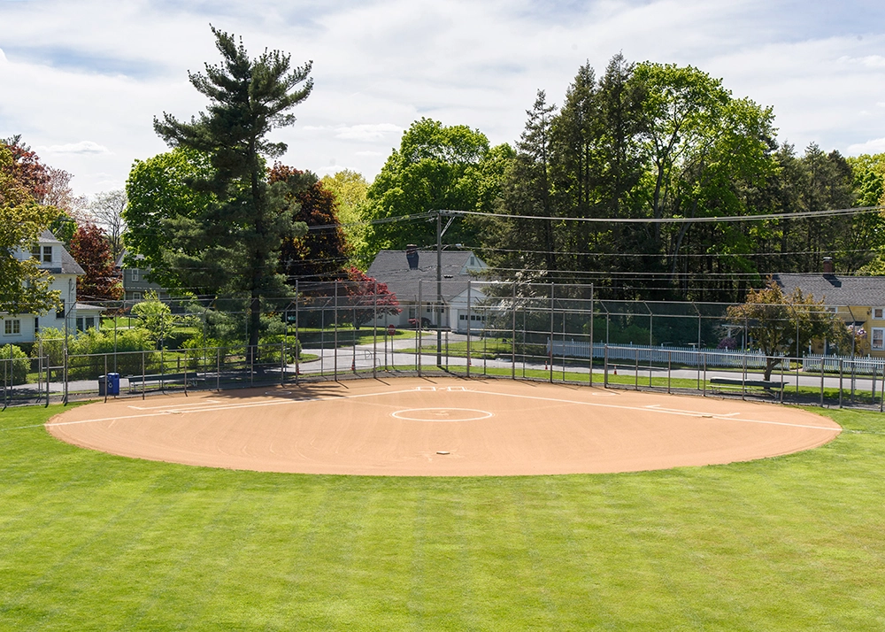 A well-maintained softball field with a dirt infield, white bases, and green grass outfield, surrounded by trees and houses, with a road and netted backstop in the background.