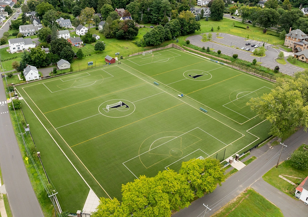 Aerial view of a large green turf sports field with multiple soccer and lacrosse markings, surrounded by trees, houses, parking lots, and nearby roads in a suburban neighborhood.