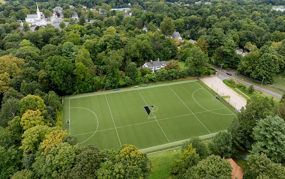 Aerial view of an empty, green athletic field with a large letter P at the center, surrounded by trees, houses, and a nearby road in a suburban neighborhood.