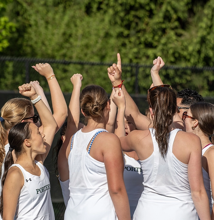 A group of girls in white athletic uniforms stand in a circle outdoors, raising their hands together in a team huddle, with greenery and a fence in the background.