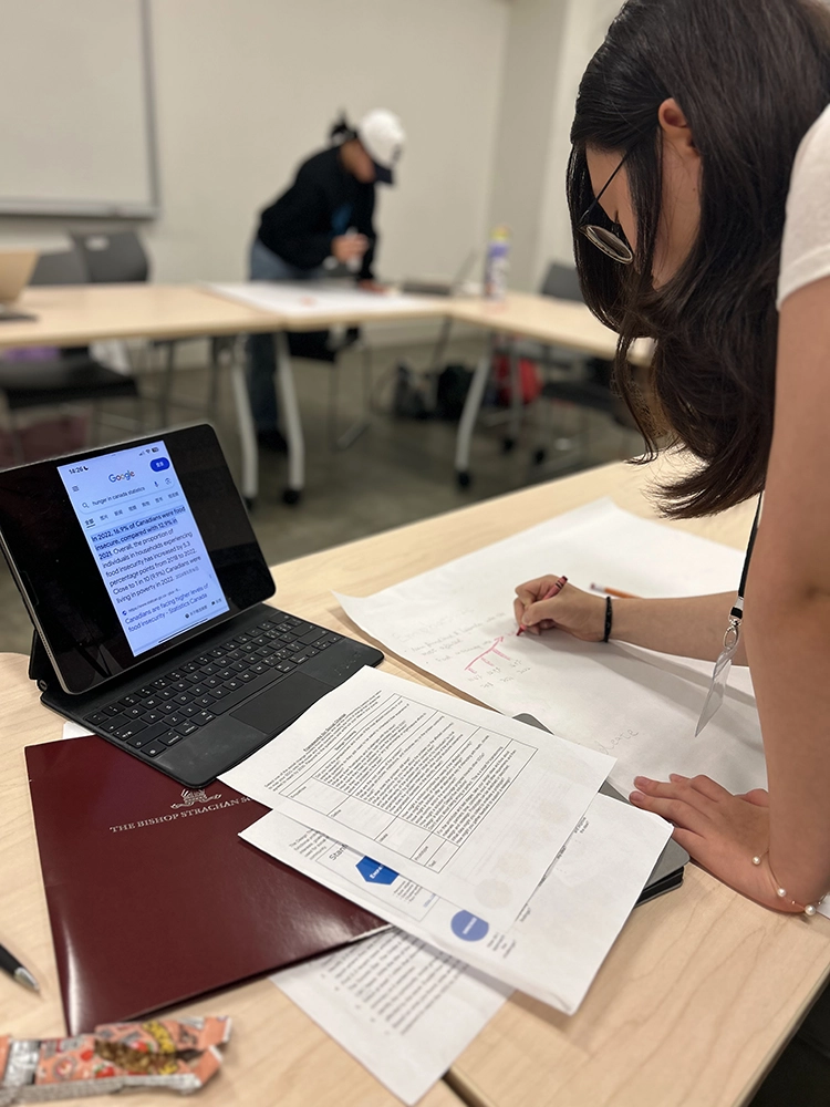 A person writes on a large sheet of paper at a table with documents and an open laptop displaying text. Another person stands in the background, also working at a table.