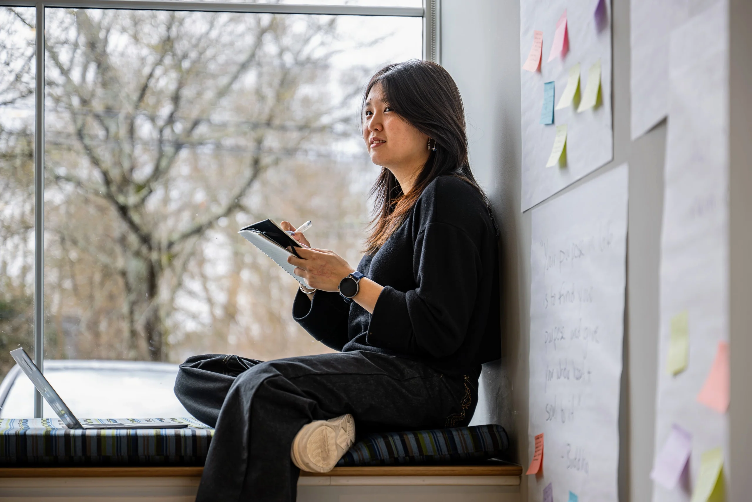 A woman sits by a window on a cushioned bench, holding a notebook and pen. She is surrounded by papers and colorful sticky notes on the wall, with a laptop nearby, appearing thoughtful and focused.