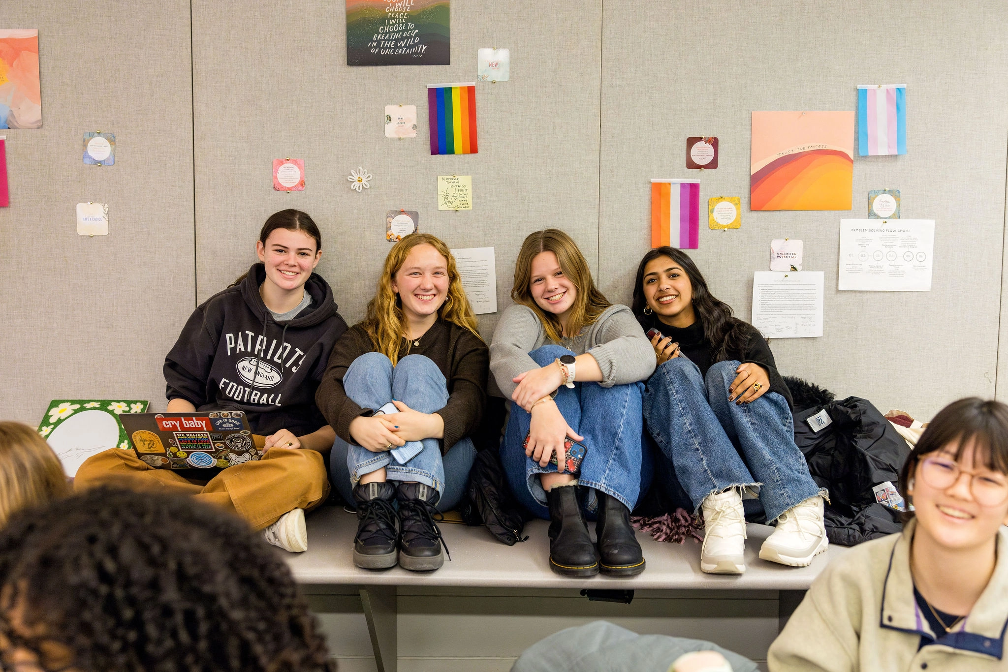 Four young people sit together on a ledge in a classroom, smiling at the camera. Colorful artwork and rainbow flags decorate the wall behind them. Other students are partially visible in the foreground.