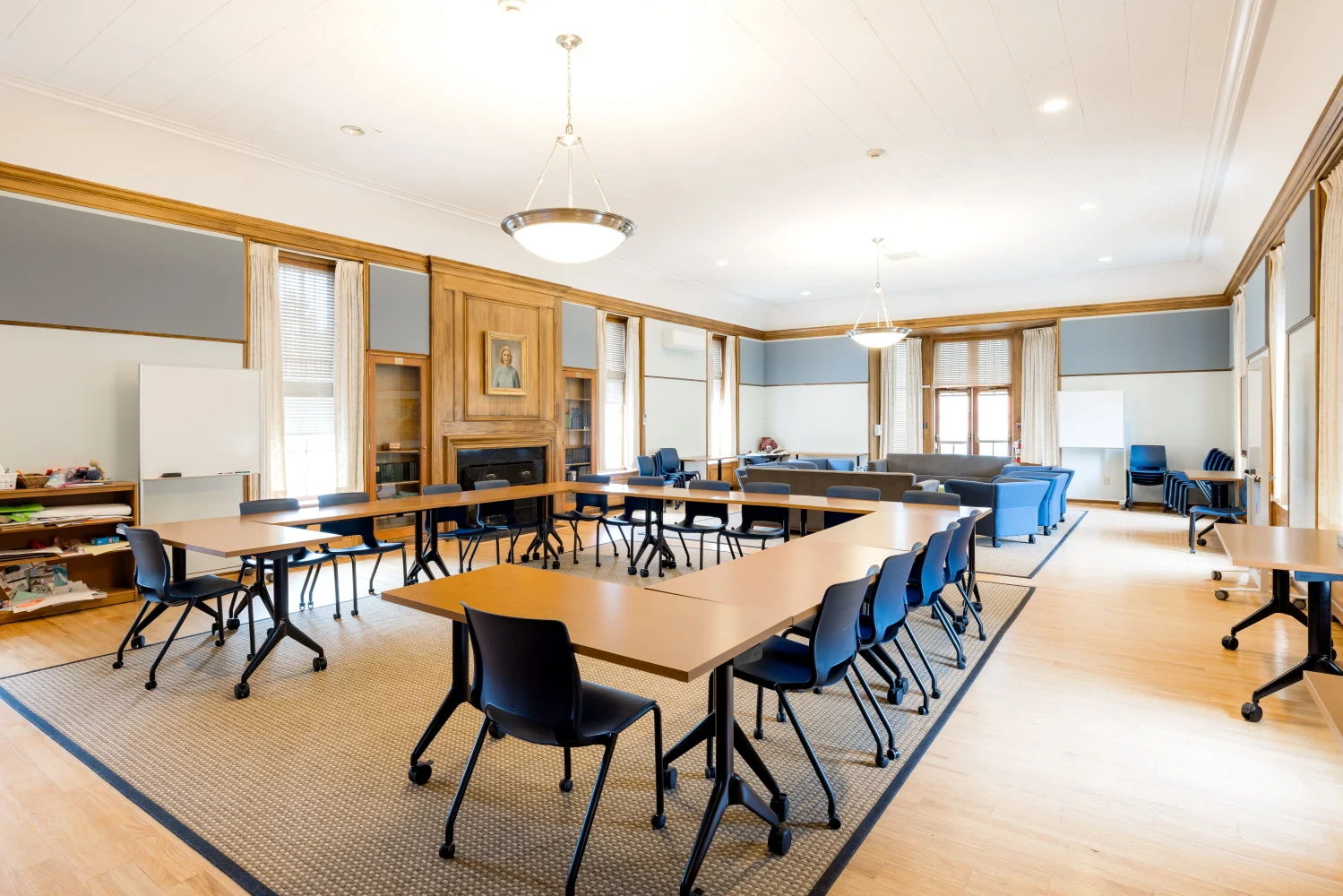 A spacious, well-lit meeting room with wooden tables and black chairs arranged in a U-shape, large windows, two sofas, whiteboards, and light wooden flooring.