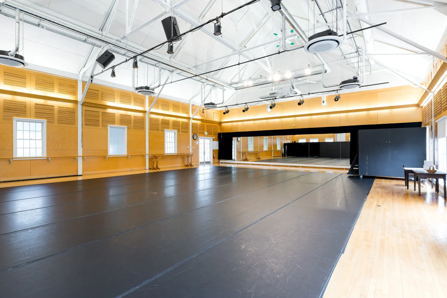 A spacious, well-lit dance studio with a black marley floor, large mirrors on one wall, ballet barres, wooden walls, and high ceilings with exposed beams and lighting fixtures.