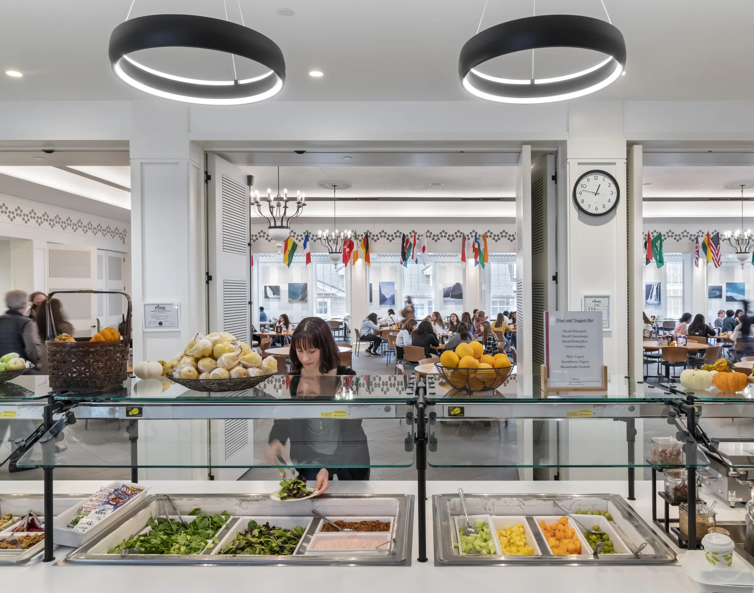 A woman prepares a salad at a self-serve salad bar in a bright cafeteria. In the background, people are dining at tables, with colorful flags and natural light visible through large windows.