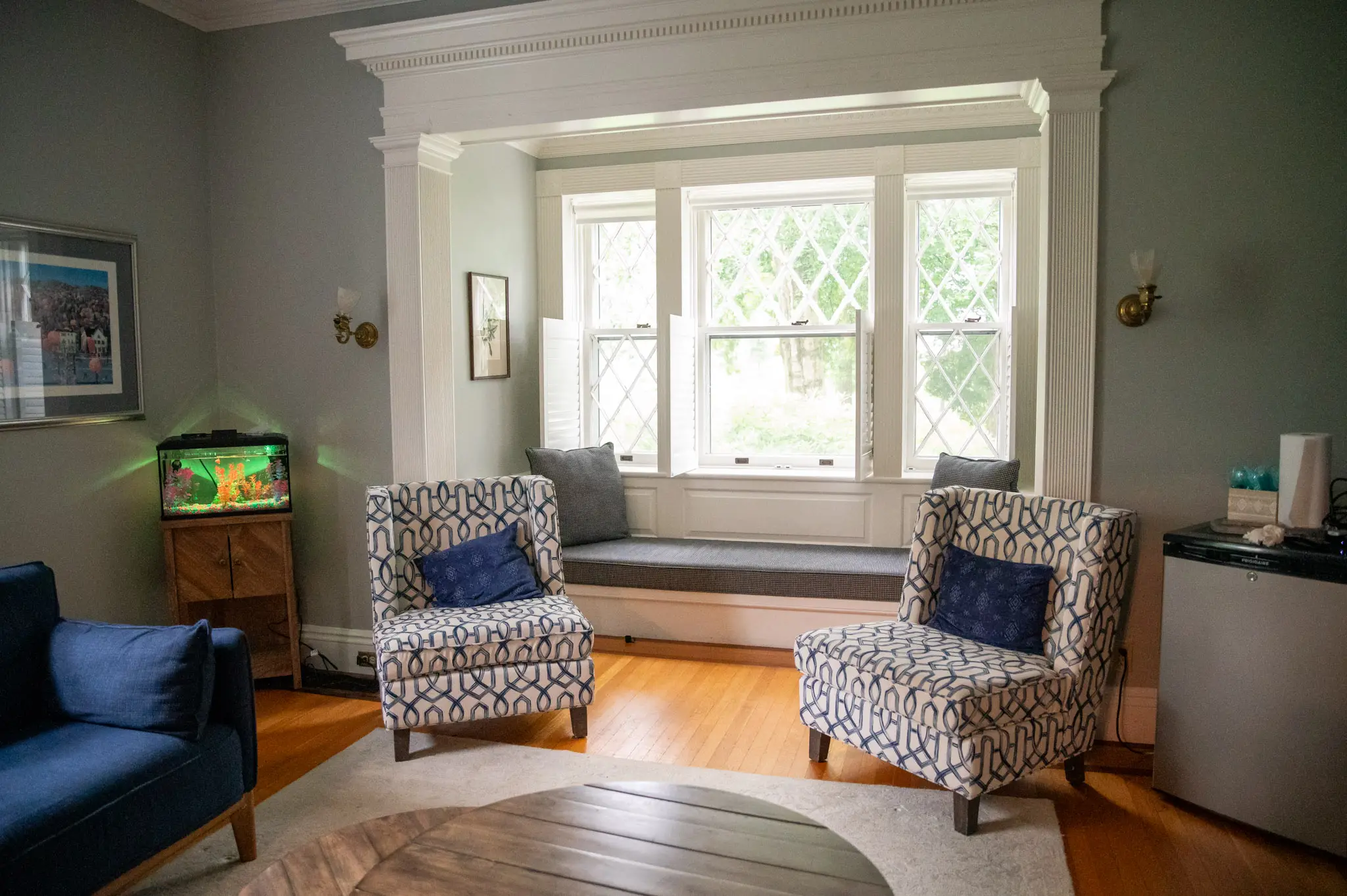 A cozy living room with two patterned armchairs, a blue sofa, a fish tank, a mini fridge, and a window seat beneath large windows letting in natural light. The room features hardwood floors and soft gray walls.