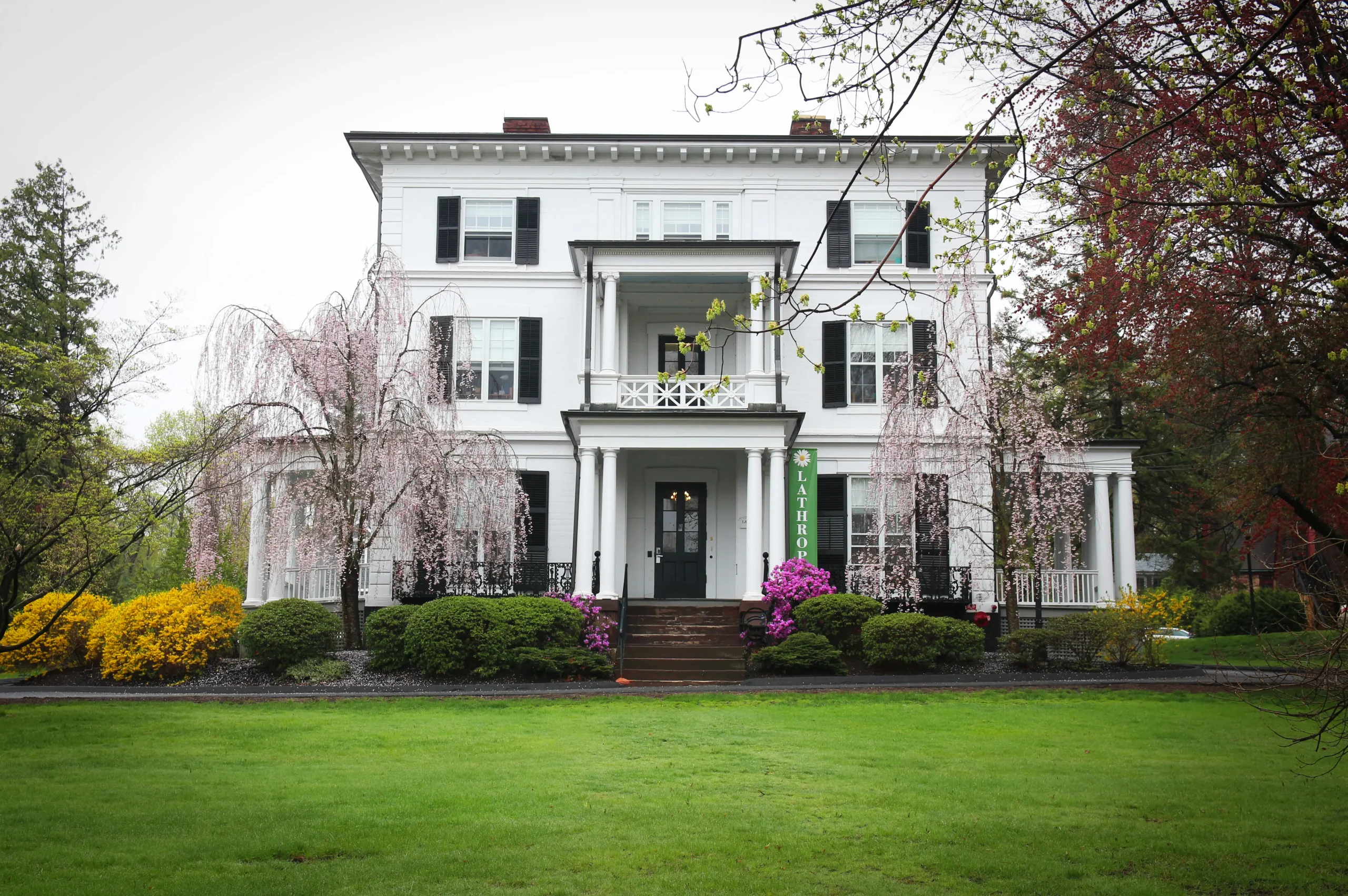 A large white two-story house with columns and a covered porch, surrounded by green lawn, blooming shrubs, and trees with pink and yellow flowers on a cloudy day.