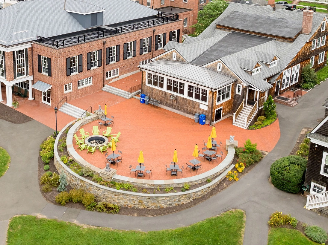 Aerial view of a brick and wood building with a circular outdoor patio, yellow umbrellas, tables, chairs, and a central fire pit, surrounded by greenery and walkways.