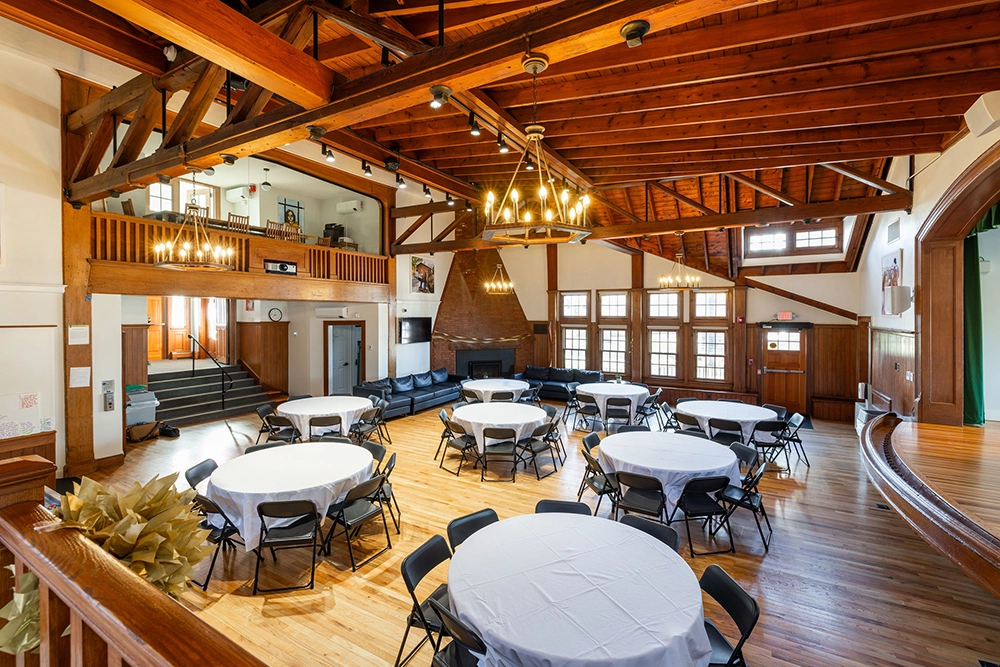 A spacious hall with exposed wooden beams, round tables covered with white cloths, black chairs, wooden floors, large windows, chandeliers, a stage area, and a lofted balcony overlooking the room.