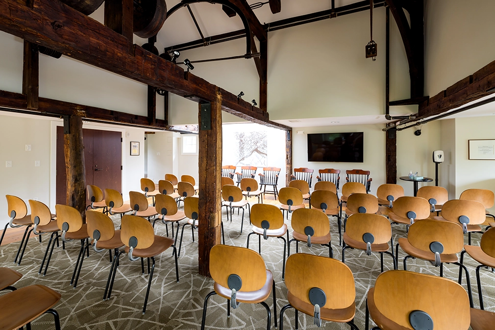 A bright conference room with rows of empty wooden chairs facing a window and a large TV screen, exposed wooden beams, and patterned carpet.