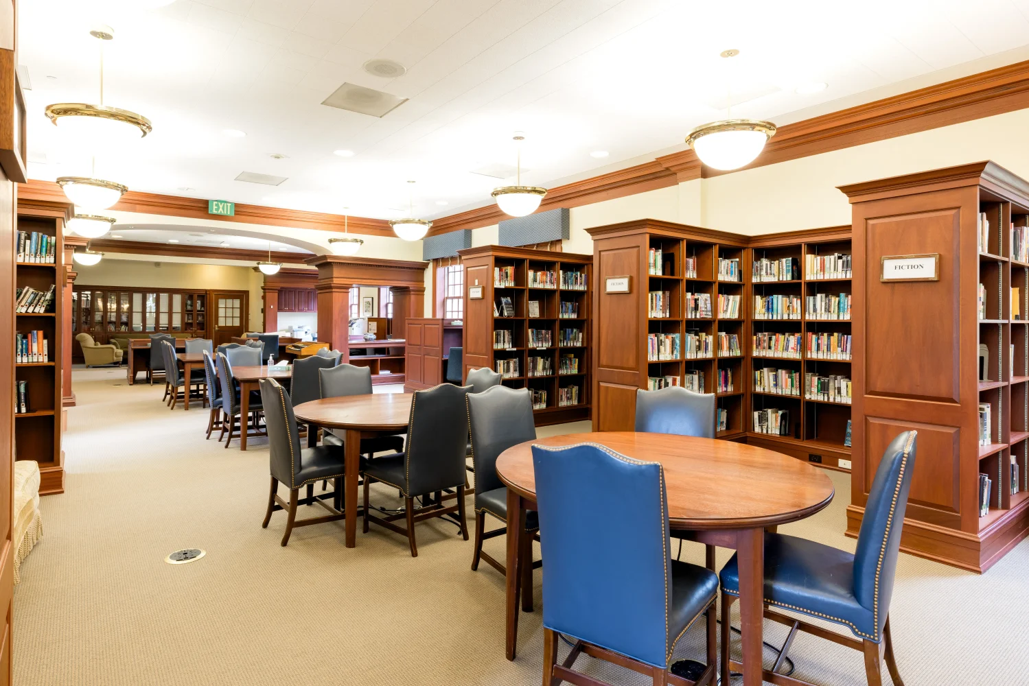 A spacious, well-lit library with wooden bookshelves filled with books, round tables surrounded by blue chairs, and large windows in the background. The room appears clean and organized.