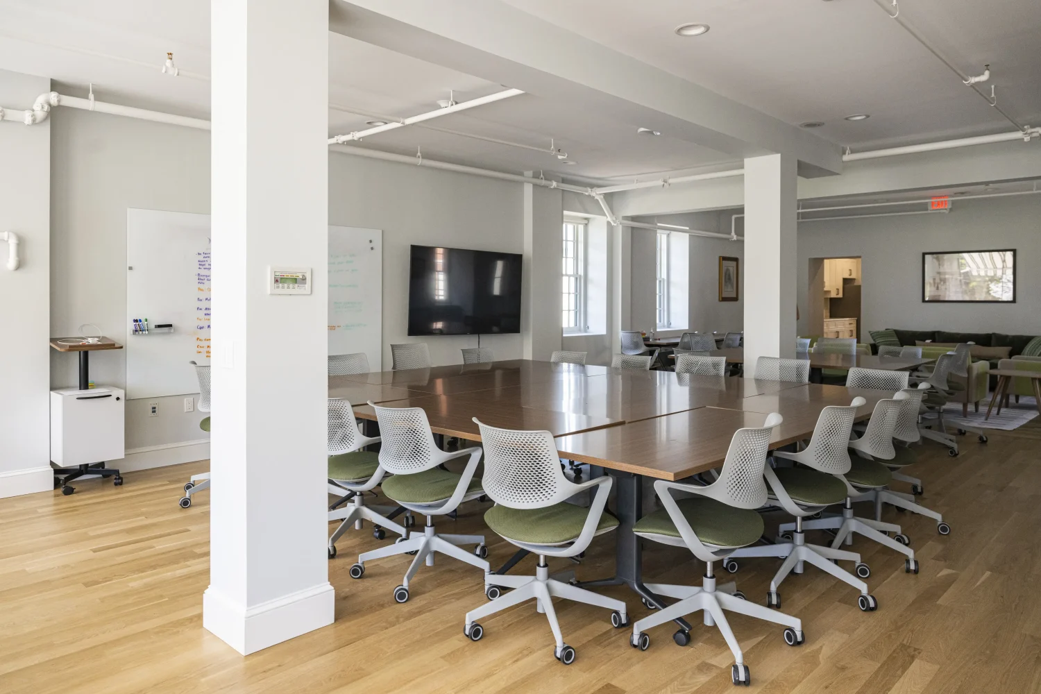 A modern conference room with a large wooden table surrounded by white office chairs on wheels, a TV mounted on the wall, a whiteboard, wood flooring, and natural light from large windows.