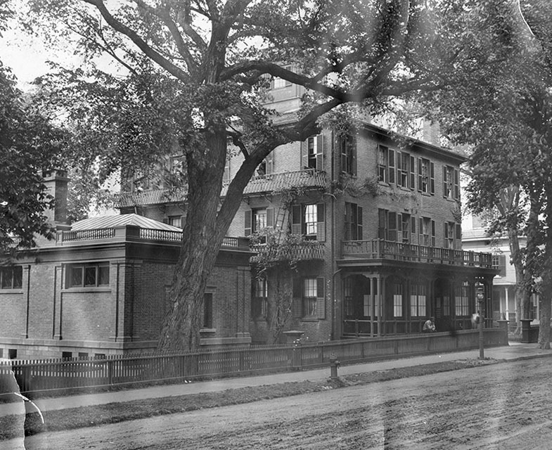 Historic black and white photo of the large large brick building that is Porter's Main, the original building of Miss Porter's School. Building has balconies and a wooden fence, shaded by tall trees. Two people stand on the porch, and a dirt road runs in front of the house.
