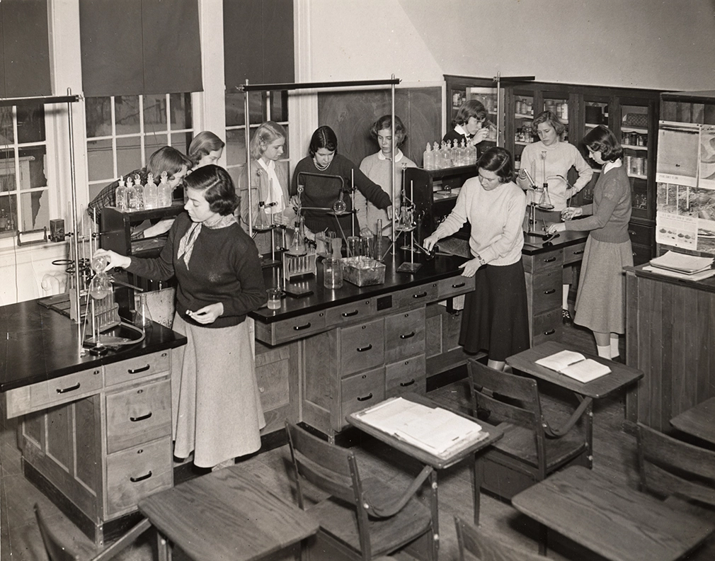 A group of women in skirts and sweaters work with lab equipment and glassware at long benches in a classroom laboratory, with shelves of bottles and books in the background. Circa 1950s.