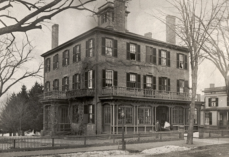 Historic black-and-white photo of a large, three-story brick house with a wraparound porch, tall chimneys, shuttered windows, bare trees, and a picket fence, taken in a winter setting.
