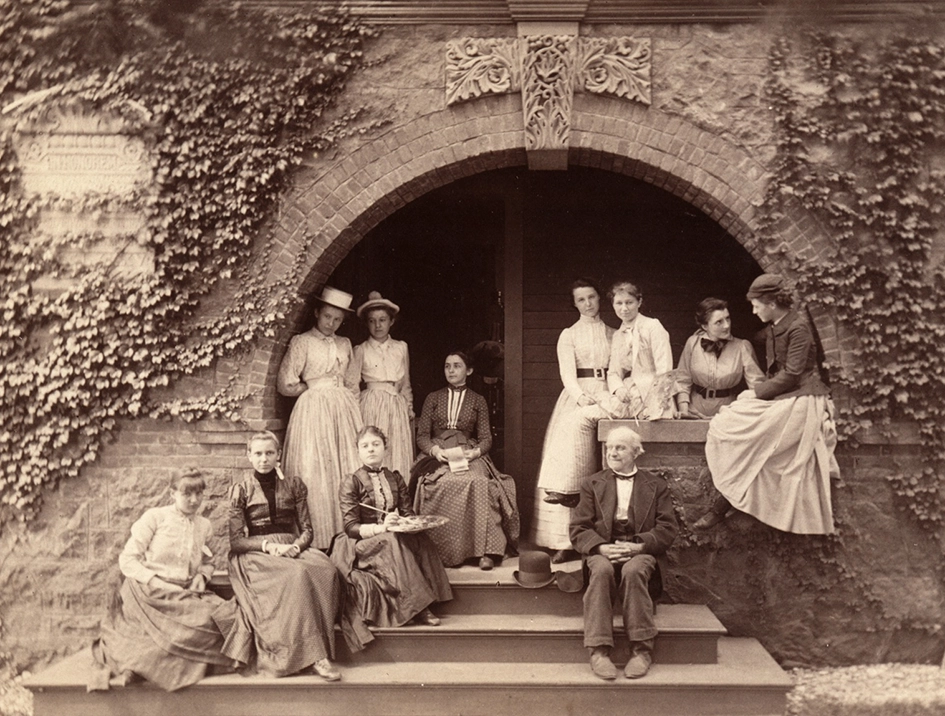 A sepia photo of nine women and one man wearing 19th-century clothing, sitting and standing on the steps of a vine-covered brick building with an arched doorway.