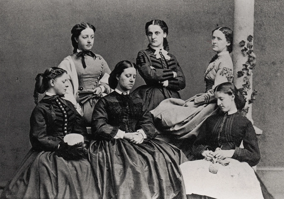 Six women in 19th-century dresses pose for a formal black-and-white portrait, some sitting, some standing, with neutral expressions against a plain backdrop and a column partially covered with ivy.