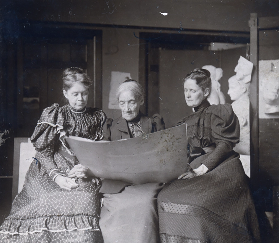Three women in Victorian-era dresses sit closely together, examining a large sheet of paper. Bust sculptures are visible in the background, suggesting an art studio or workshop setting.