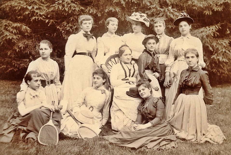 A sepia-toned vintage photo of thirteen women in Victorian-era dresses, some seated and some standing, outdoors on grass with trees in the background. Three women in front hold tennis rackets.