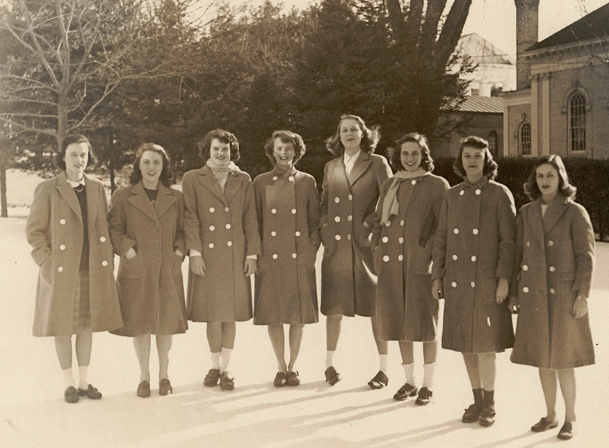 Seven young women stand outdoors in winter coats, smiling, with trees and a building in the background. The photo appears vintage, possibly from the 1940s or 1950s.