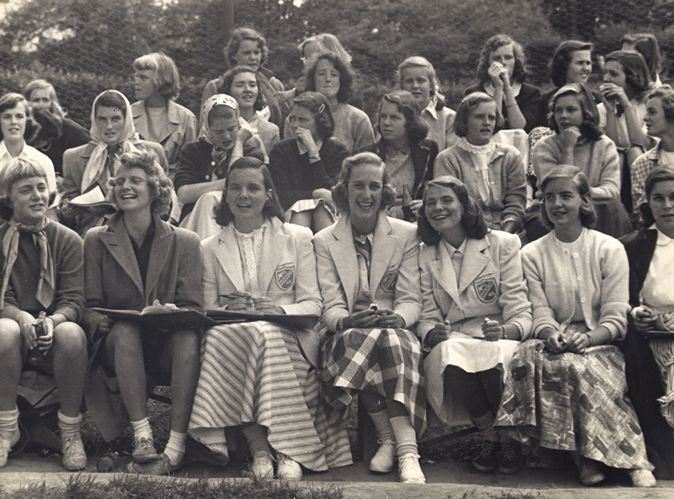 A group of young women, dressed in skirts, blouses, and sweaters, sit closely together on bleachers outdoors, smiling and talking, in a black-and-white vintage photograph. Trees and a fence are visible in the background.