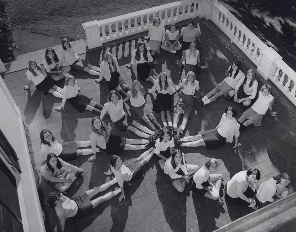 Black and white photo of young women seated in a circular formation on a tiled terrace, with their legs extended radially outward, creating a flower-like pattern. They are dressed in shirts and skirts.
