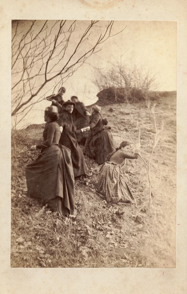Six women in long dresses are climbing up a grassy, leaf-covered hill. Bare tree branches and shrubs are visible, suggesting a late autumn or winter setting. The scene has an old, sepia-toned photograph style.