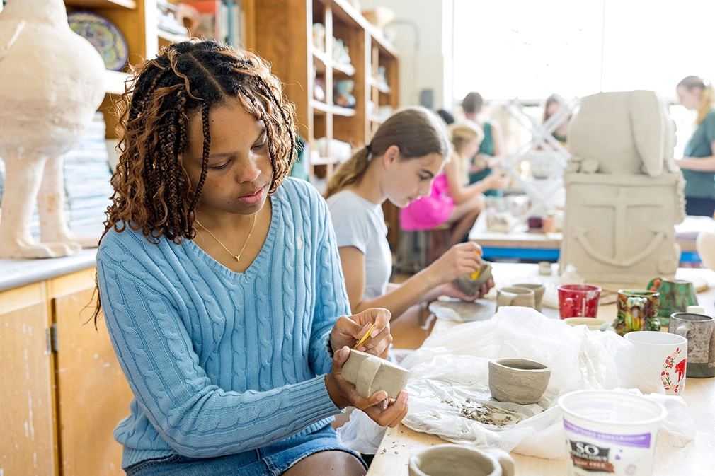 A woman in a blue sweater shapes clay with a tool in a pottery classroom, while other students in the background work on their own clay projects at a shared table.