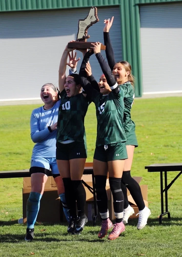 Four young women in soccer uniforms celebrate on grass, holding a trophy overhead. Two players in green jerseys lift the trophy, flanked by a teammate in blue and another in green, all smiling and cheering by a green building.