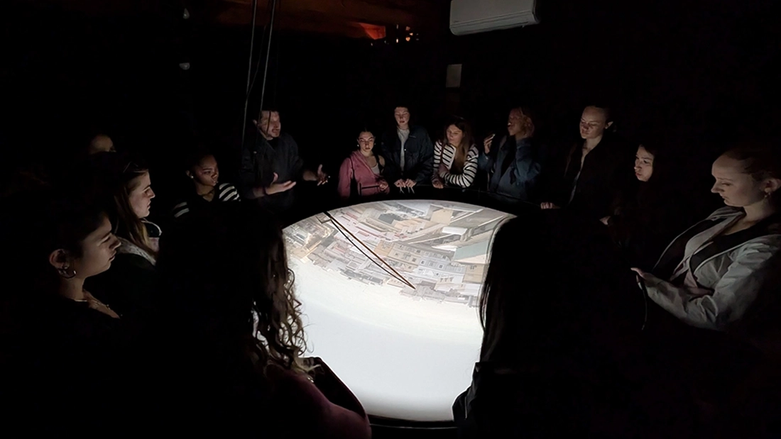 A group of people stand in a dark room around a circular table, viewing an upside-down projected image of a cityscape in the center of the table.