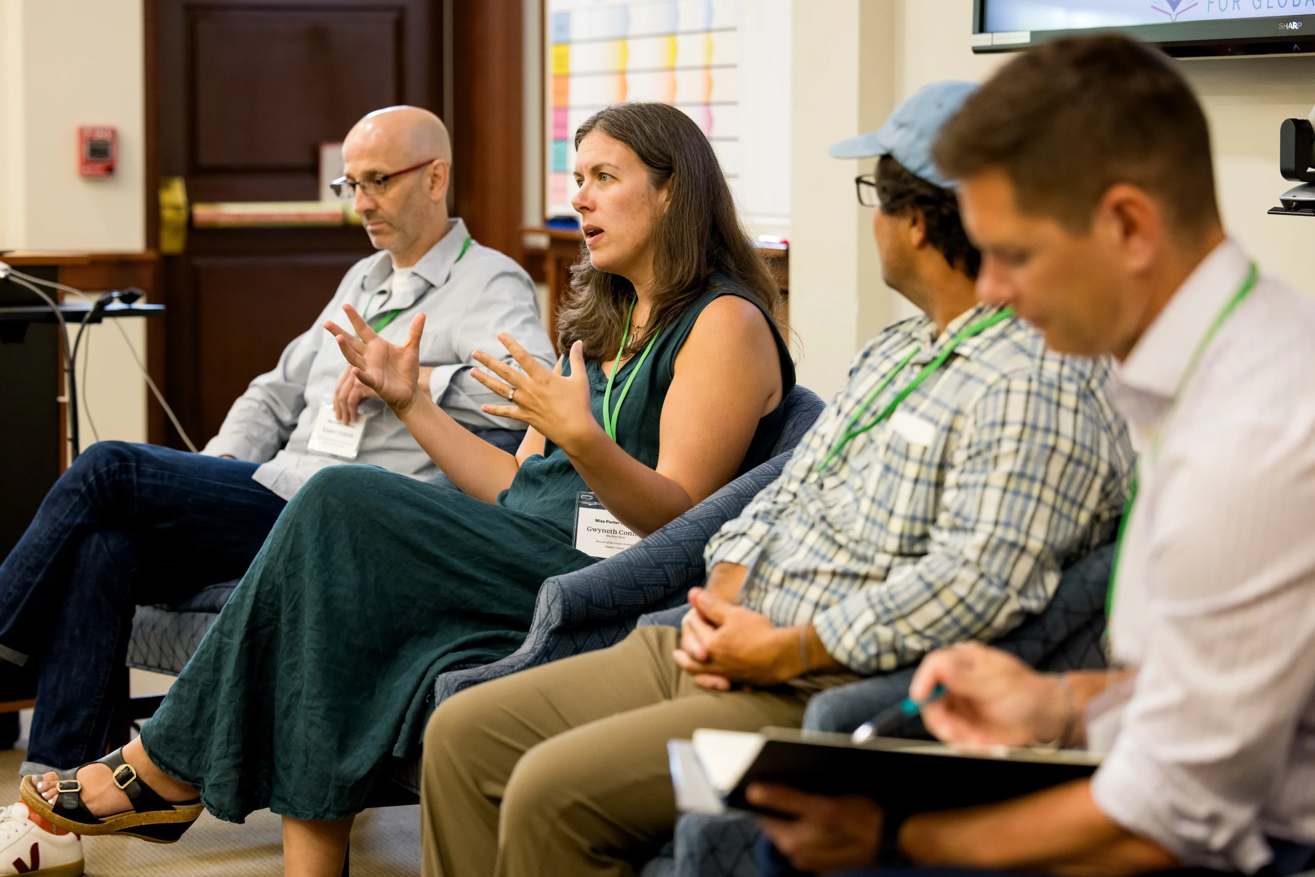 Four people sit in a panel discussion for an education professional development workshop; one woman gestures while speaking, and the others listen. They all have name tags and green lanyards. One man in front is writing in a notebook.