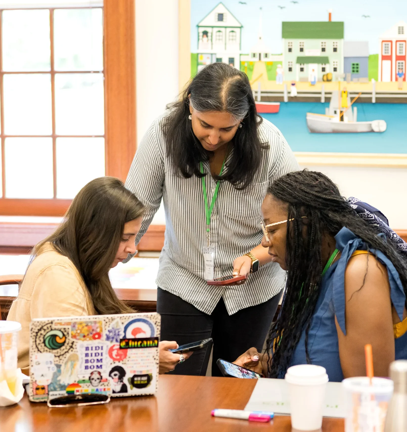Three women gather around a table with laptops, drinks, and notebooks, collaborating on a project for teaching professional development in a brightly lit room with a colorful painting and large windows in the background.
