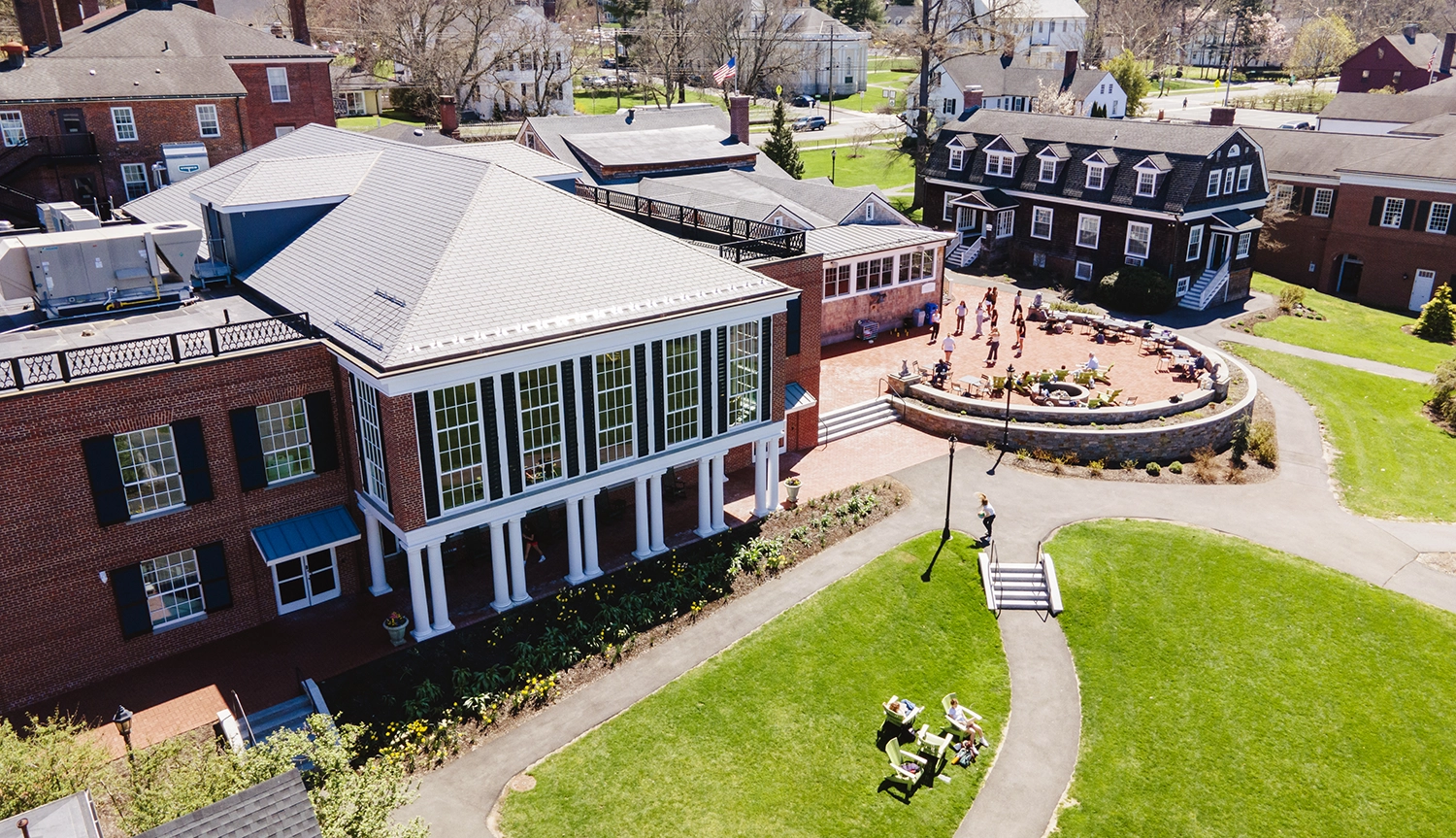 Aerial view of MPS center campus with a red-brick building with tall windows and columns, adjacent to a courtyard where people sit at round tables. Surrounding lawn and pathways are visible under a clear sky.
