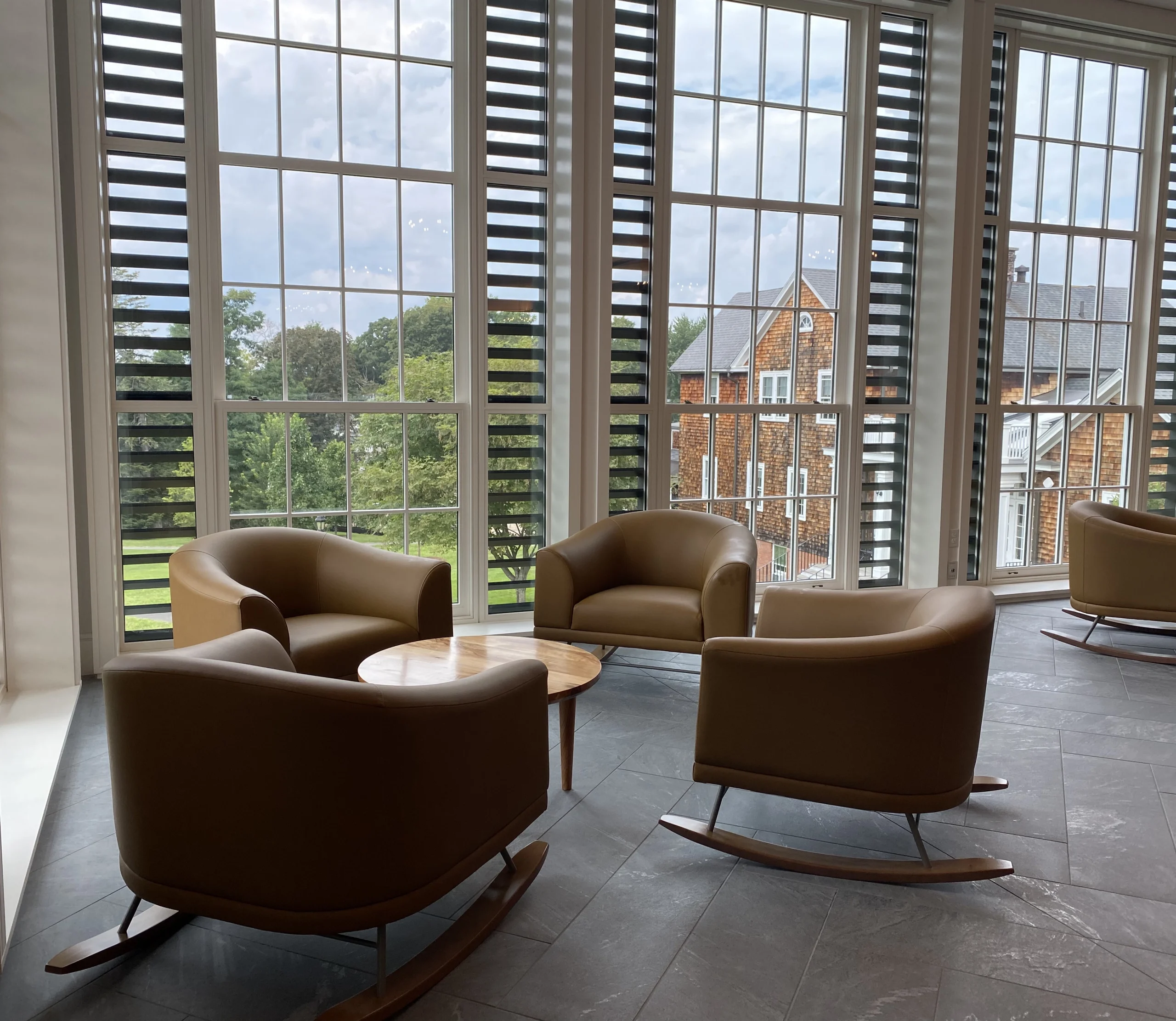 Four tan armchairs arranged around a small round wooden table in front of large windows with a view of trees and a brick building outside. The chairs sit on a gray tiled floor in a bright, modern room.