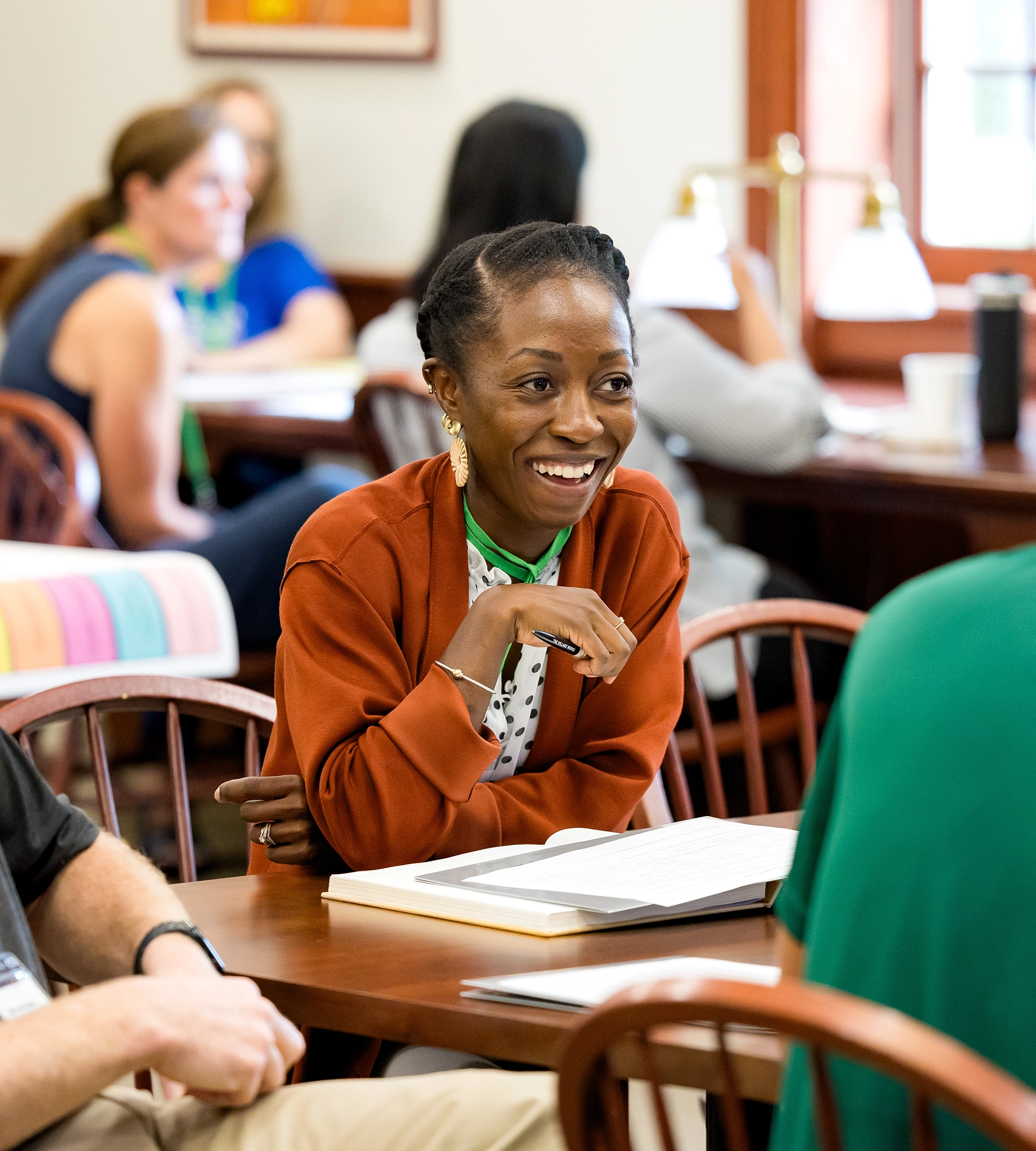 A woman sits at a table in a sunny room, smiling and engaging in conversation about opportunities for educators. She wears a rust-colored jacket and has a book open in front of her, as others chat in the background.