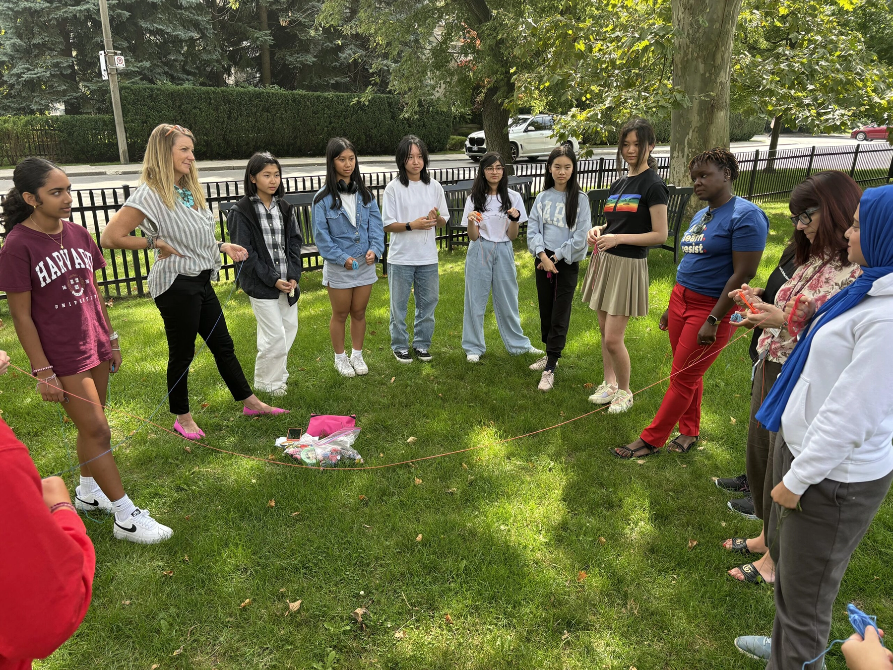 A diverse group of people stand in a circle on grass, holding a long string for a team building activity. Some are smiling and interacting while sunlight filters through the trees. There are objects on the ground in the center of the circle.