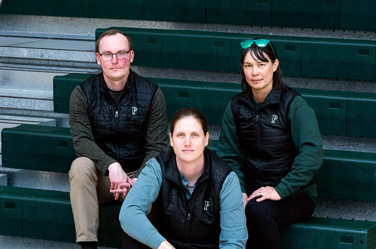 Three adults in athletic wear sit on gym bleachers; two sit on the bench, one sits below with a basketball in front. All have serious expressions. The bleachers and gym floor are green and wooden.
