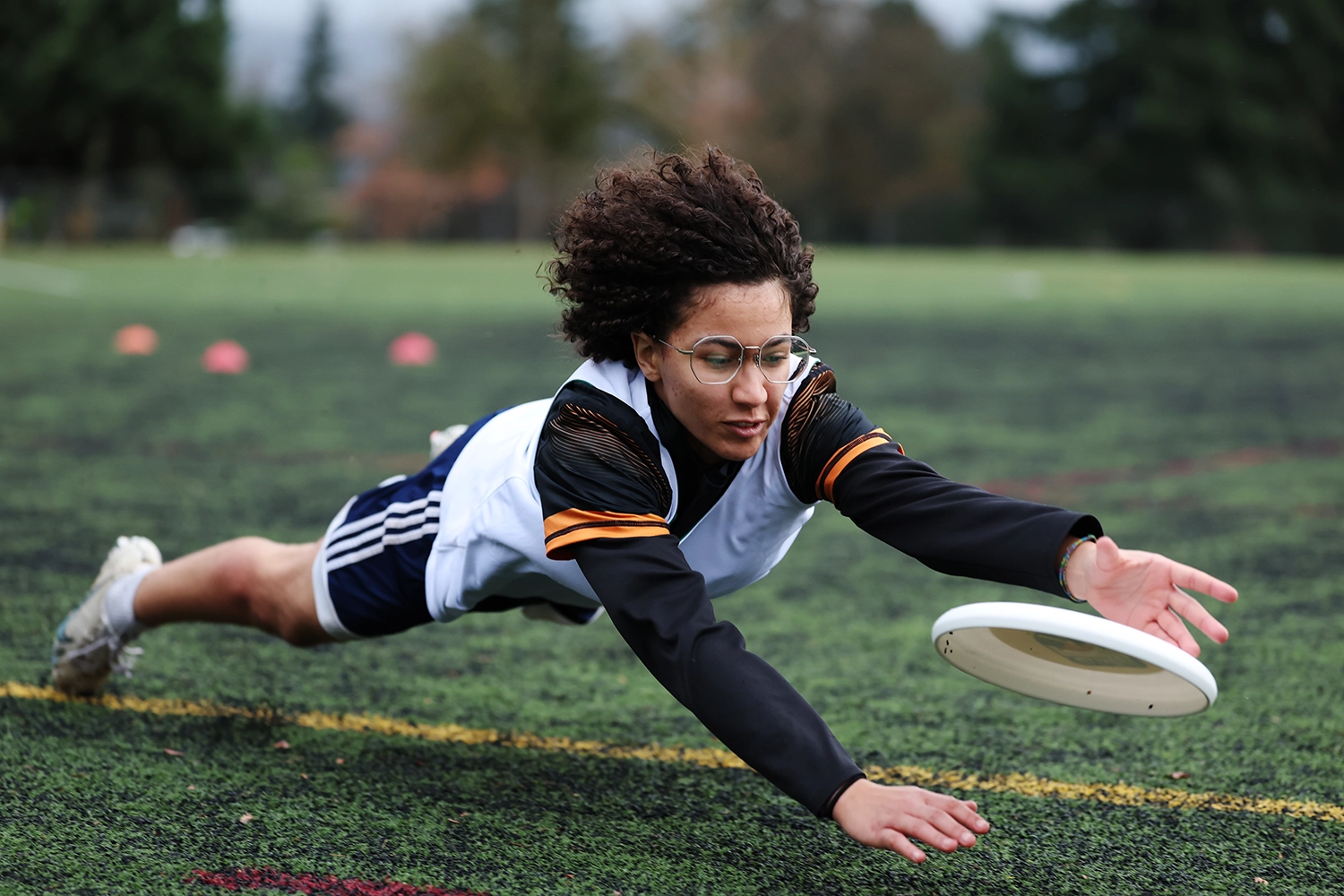 A person wearing glasses and sports gear dives on a grassy field to catch a flying white frisbee, with trees and cones blurred in the background.