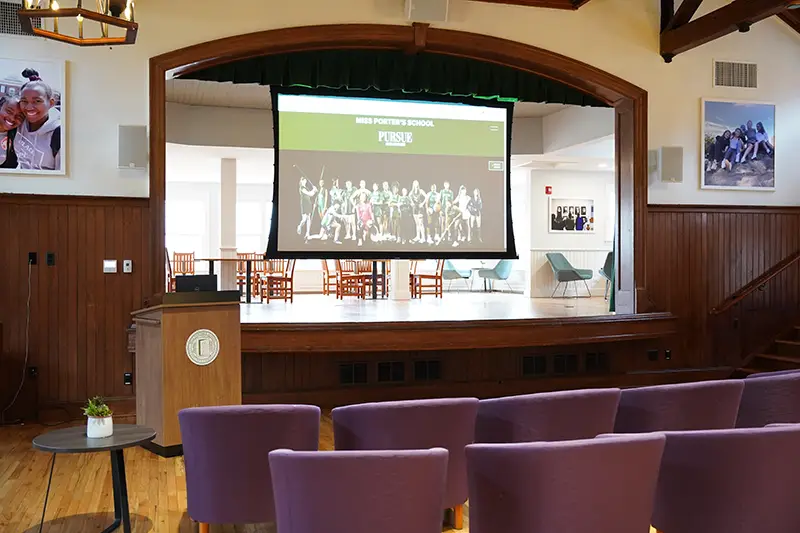 A small auditorium with purple chairs faces a stage and projection screen displaying Miss Porters School. A podium stands to the left; framed photos and a window are on the wood-paneled walls.