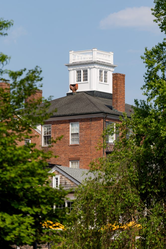 A red brick building with a white cupola on the roof is partially obscured by green trees, under a blue sky with a few clouds.