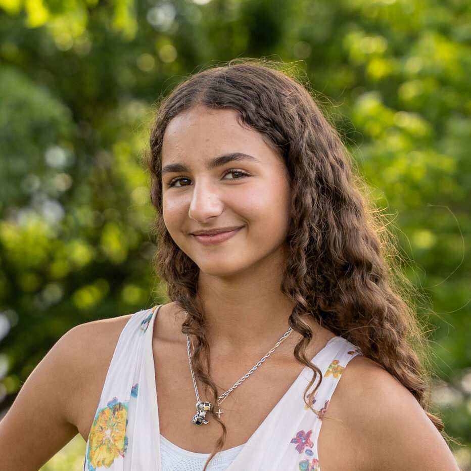 A young woman with long curly hair, wearing a sleeveless white floral dress, stands outdoors with her hands on her hips, smiling. The background is lush and green with trees and sunlight.