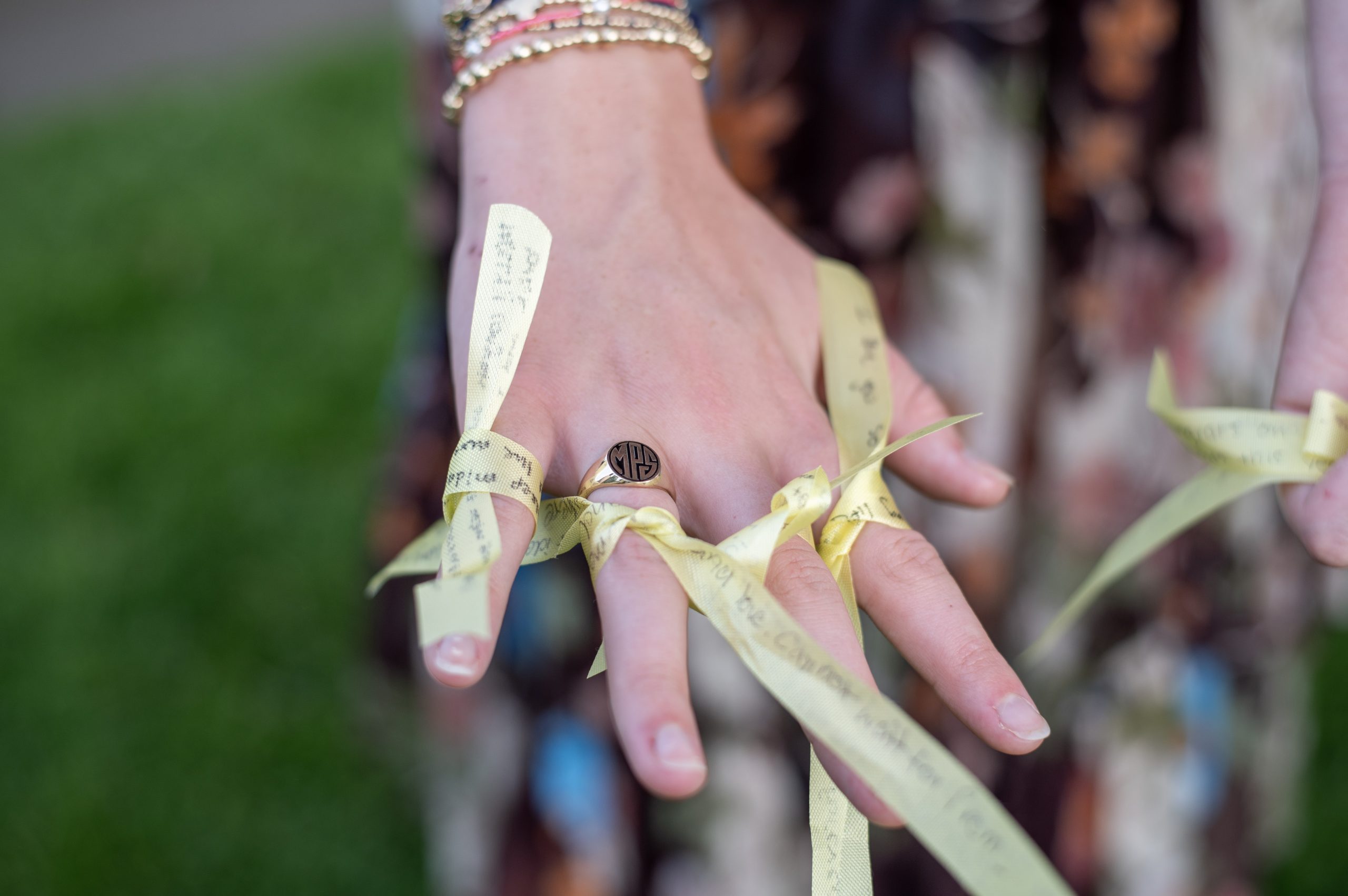 A close-up of a hand with a ring on the middle finger, adorned with yellow ribbons tied around the fingers. The background is blurred grass and patterned clothing.
