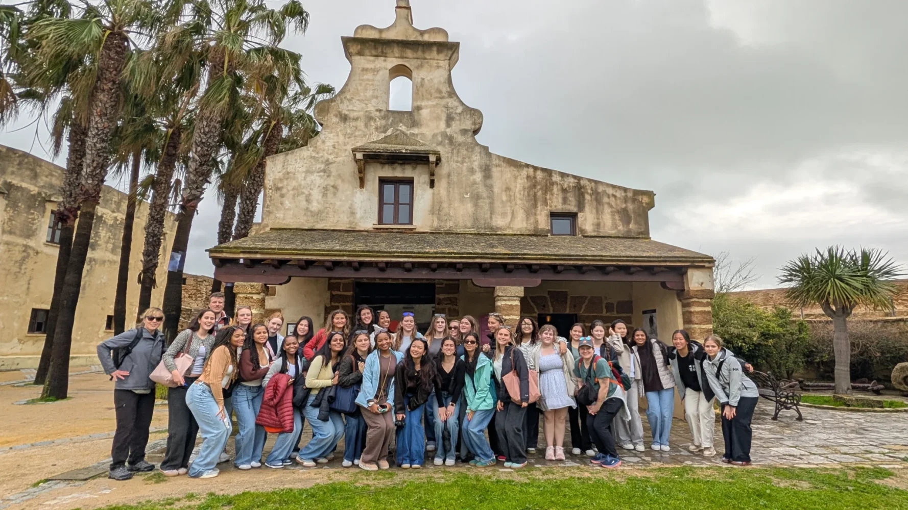 A large group of people poses and smiles in front of an old stone building with a bell tower, under a cloudy sky. There are palm trees nearby and grass in the foreground.