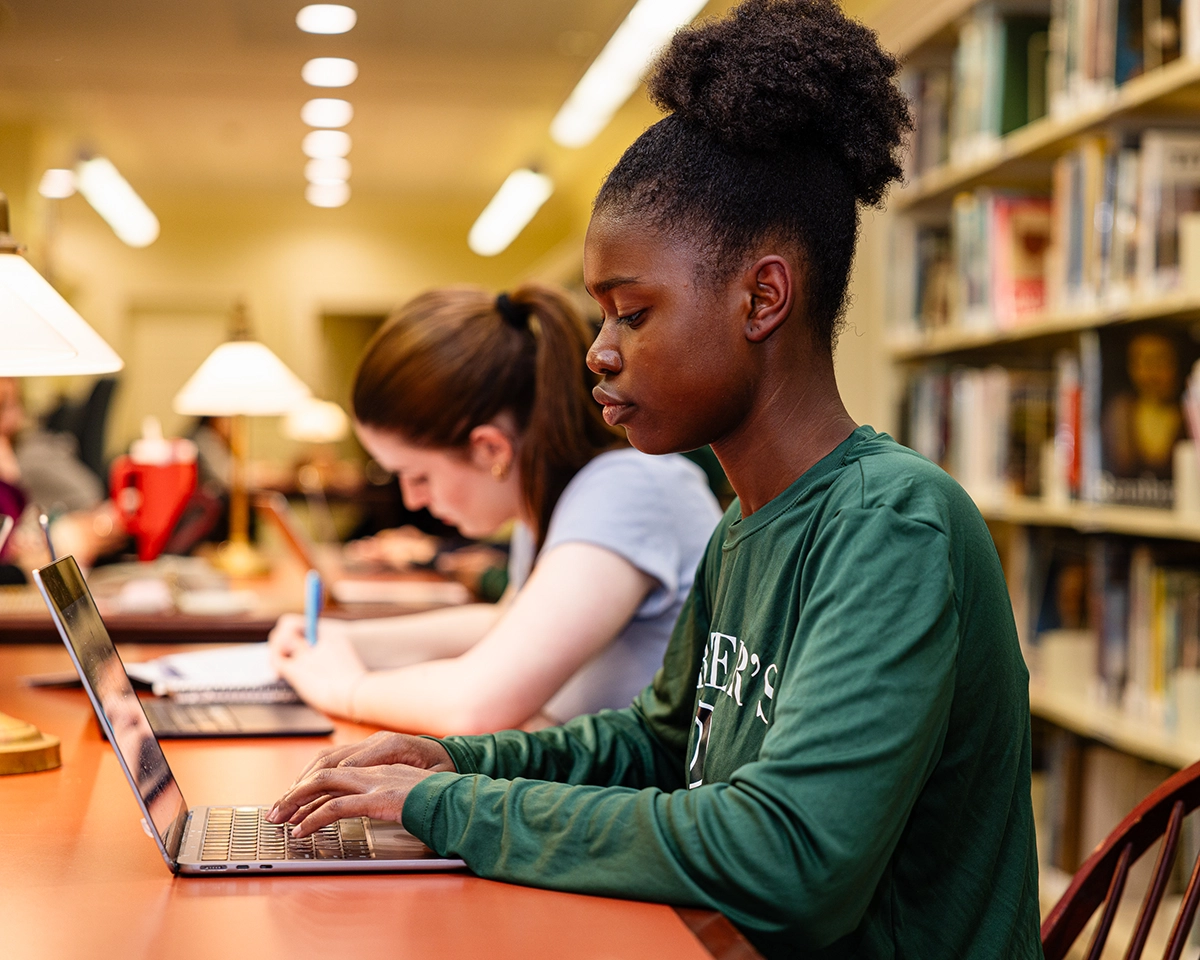 Two students sit at a library table, working. The student in front types on a laptop, while the student behind writes in a notebook. Bookshelves and lamps are visible in the background.