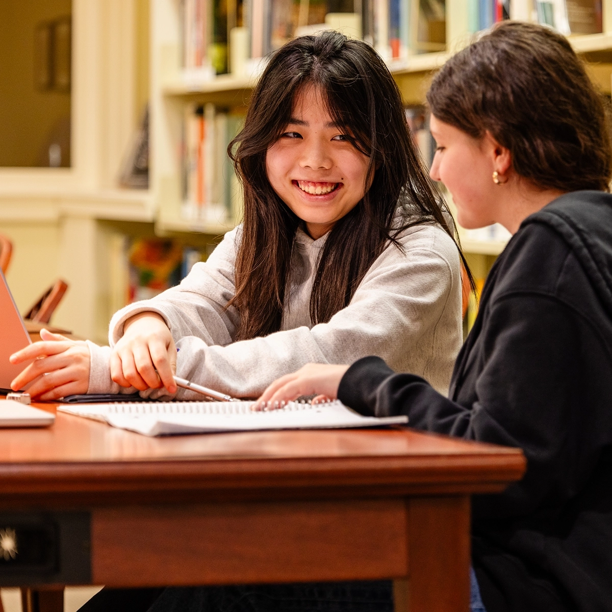 Two students sit at a library table, smiling and talking while studying together. One points at a notebook; bookshelves are visible in the background.