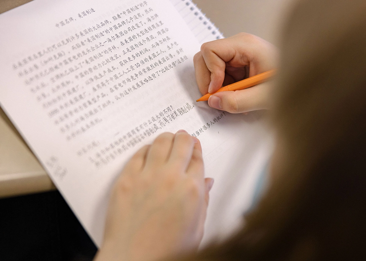 A person writes with an orange pencil on a notebook filled with handwritten Chinese text. Only their hands and a partial notebook are visible.