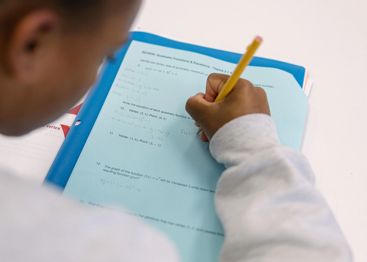 A student writes with a pencil on a blue worksheet filled with math equations, viewed from above. The students hand and part of their arm are visible, while their face is out of focus.